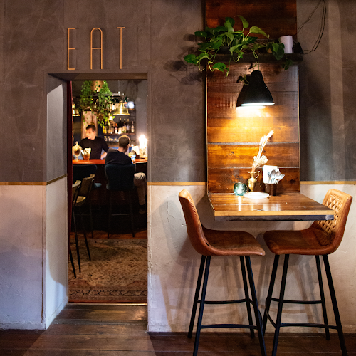 Dimly lit interior at Night Kitchen Amsterdam, with brass EAT lettering above a doorway into the bar room and two tan leather stools at a high table under a black pendant lamp