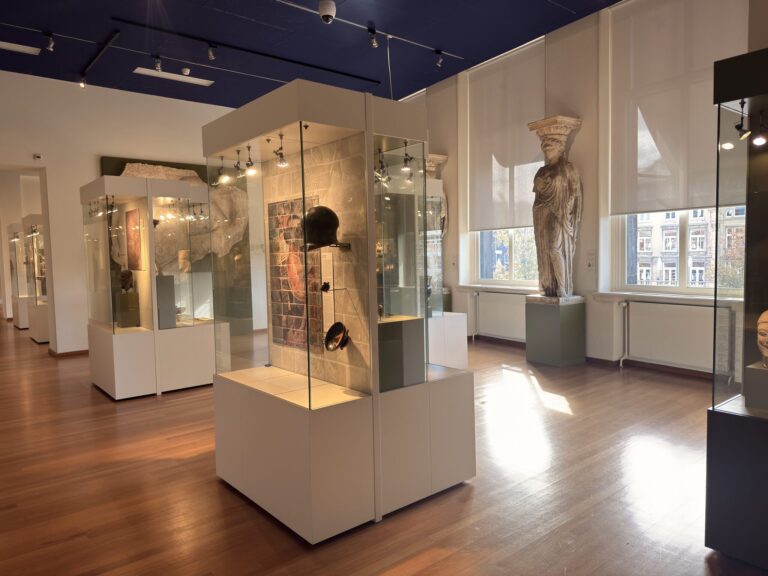 Sunlit Greek antiquities gallery with a Caryatid statue cast by the windows and glass display cases of ancient armor