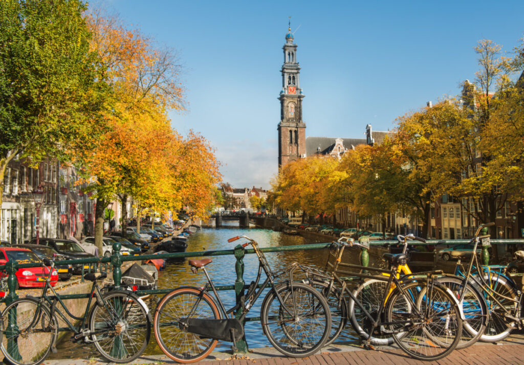 10 Pin‑Drop Perfect Places for Fall Colors in Amsterdam (Exact Spots, Zero Guesswork) 14 Bicycles line a canal bridge in Amsterdam, framed by autumnal trees. The Westerkerk church tower rises in the background under a clear blue sky.