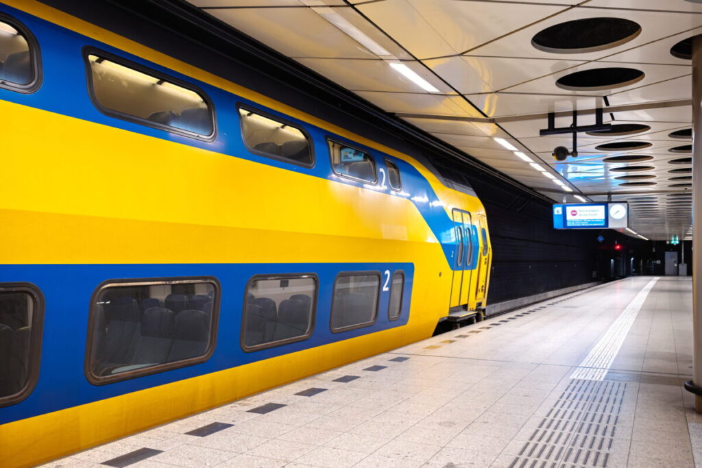 Amsterdam Airport Train vs Taxi: Which Is Better in 2026? 2 Yellow NS train stopped at Schiphol Airport railway station beneath the terminal