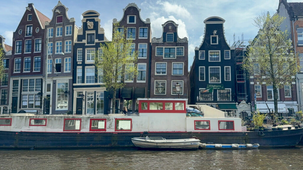 The Dancing Houses 5 Leaning Amsterdam canal houses with mismatched gables reflected in the water, a tour boat passing in front.