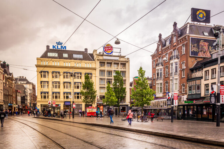 Leidseplein urban square on a rainy day, featuring colorful buildings with signs, tram tracks, scattered pedestrians, and wet streets reflecting city lights.
