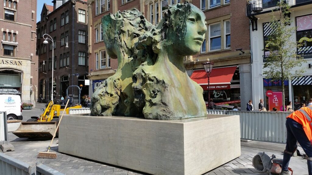 Statue of two women on a pedestal, located at Rokin Fontein in Amsterdam, surrounded by urban scenery.
