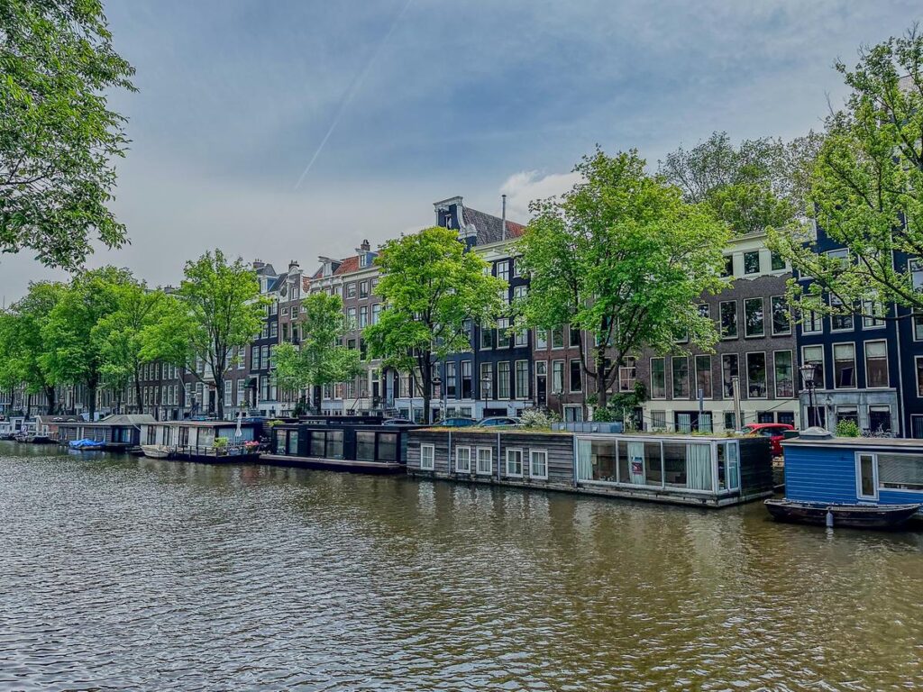 15 Iconic Amsterdam Canals: Explore the UNESCO World Heritage Waterways 5 Historic Dutch houses and houseboats along the Singel canal in Amsterdam, embraced by lush foliage and a tranquil watery expanse, under a soft spring sky.