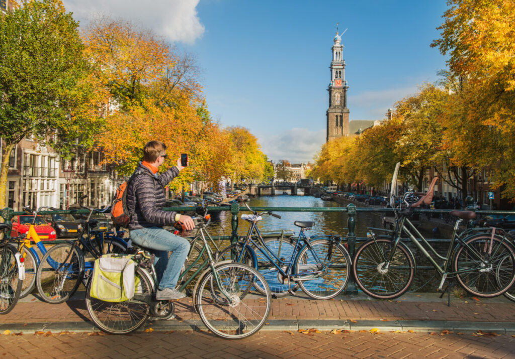 10 Pin‑Drop Perfect Places for Fall Colors in Amsterdam (Exact Spots, Zero Guesswork) 2 Man on bicycle takes photo by Amsterdam canal with autumn trees and Westerkerk tower in background